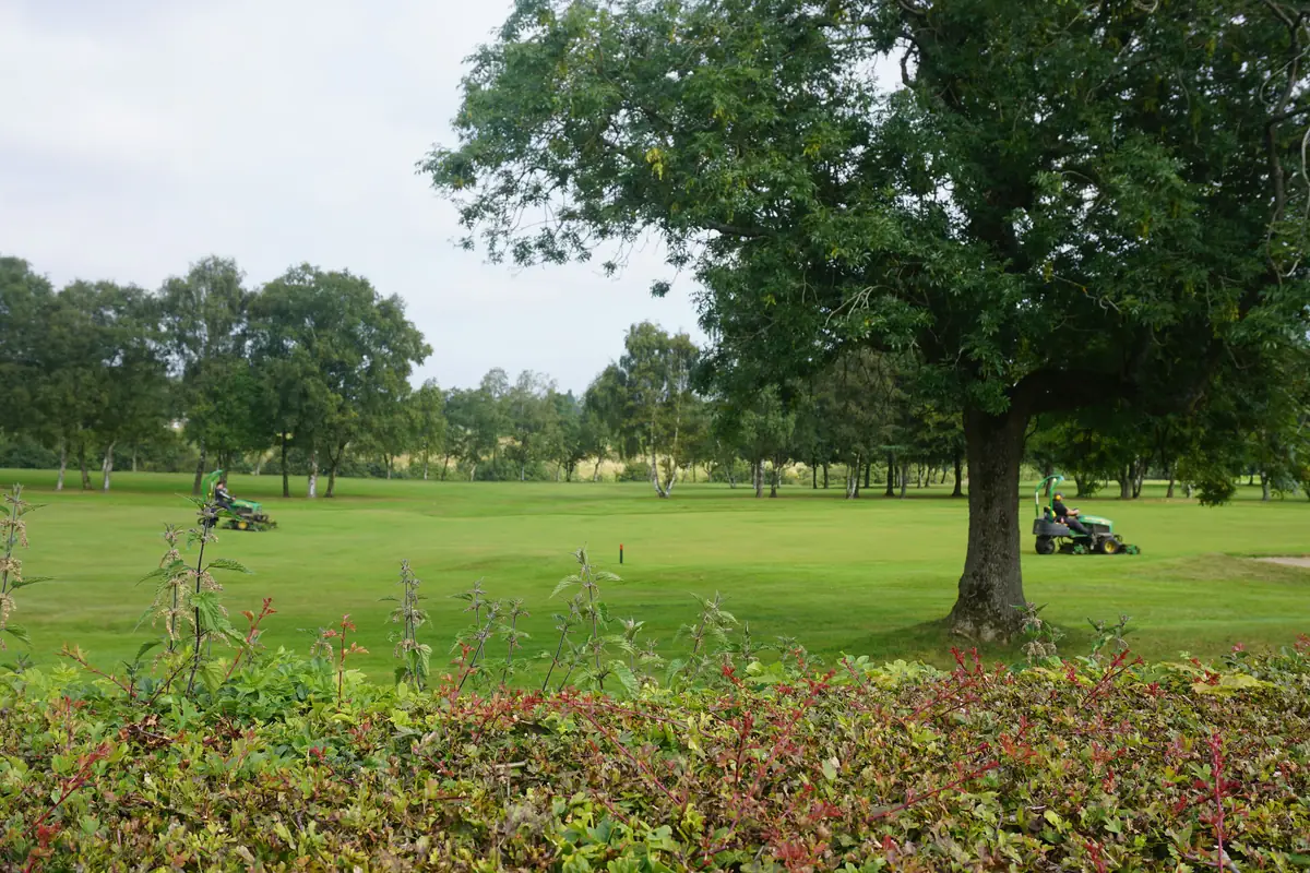 Two greenkeepers on ride-on mowers cutting the fairway at a tree-lined parkland golf course in England